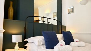 Una cama bien tendida con sábanas blancas y almohadas azules, una mesita de noche con una lámpara, y un espejo colgado en la pared.