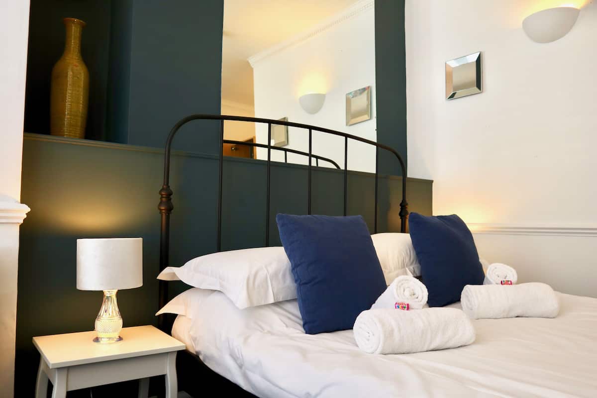 A neatly made bed with white linens and blue pillows, a bedside table with a lamp, and a wall-mounted mirror.