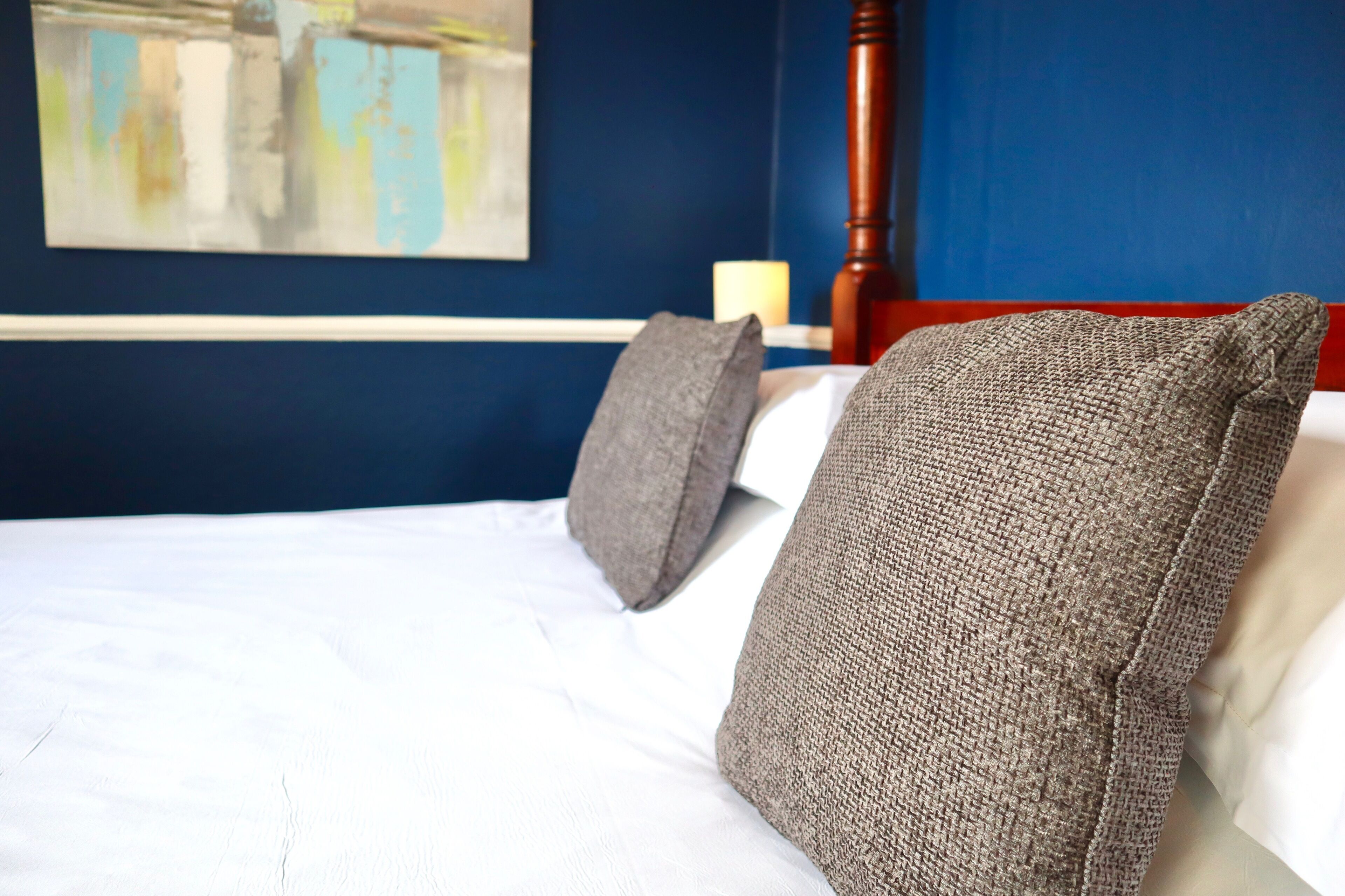 Una cama bien hecha con sábanas blancas y dos almohadas grises, un cabecero de madera, una pared azul y un cuadro sobre la cama.