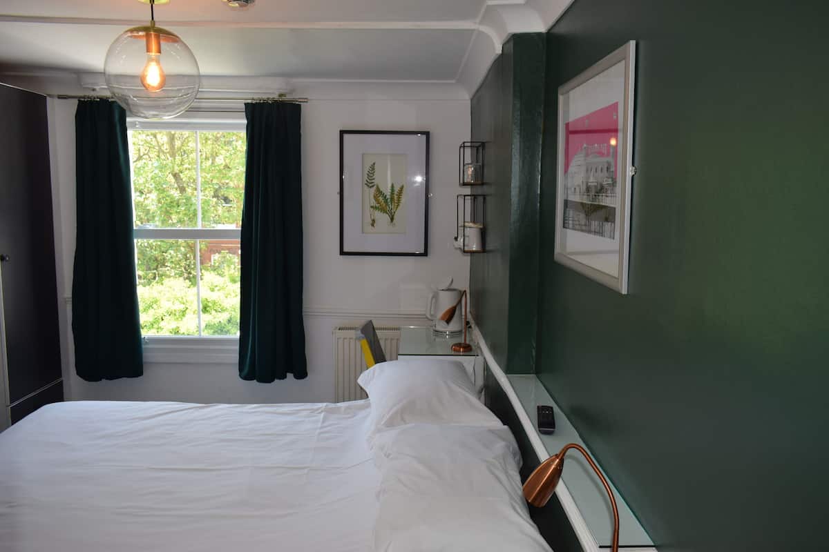 A bedroom with a bed, a window with curtains, a framed plant, a radiator, and a bedside table with a lamp.