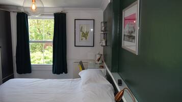 A bedroom with a bed, a window with curtains, a framed plant, a radiator, and a bedside table with a lamp.