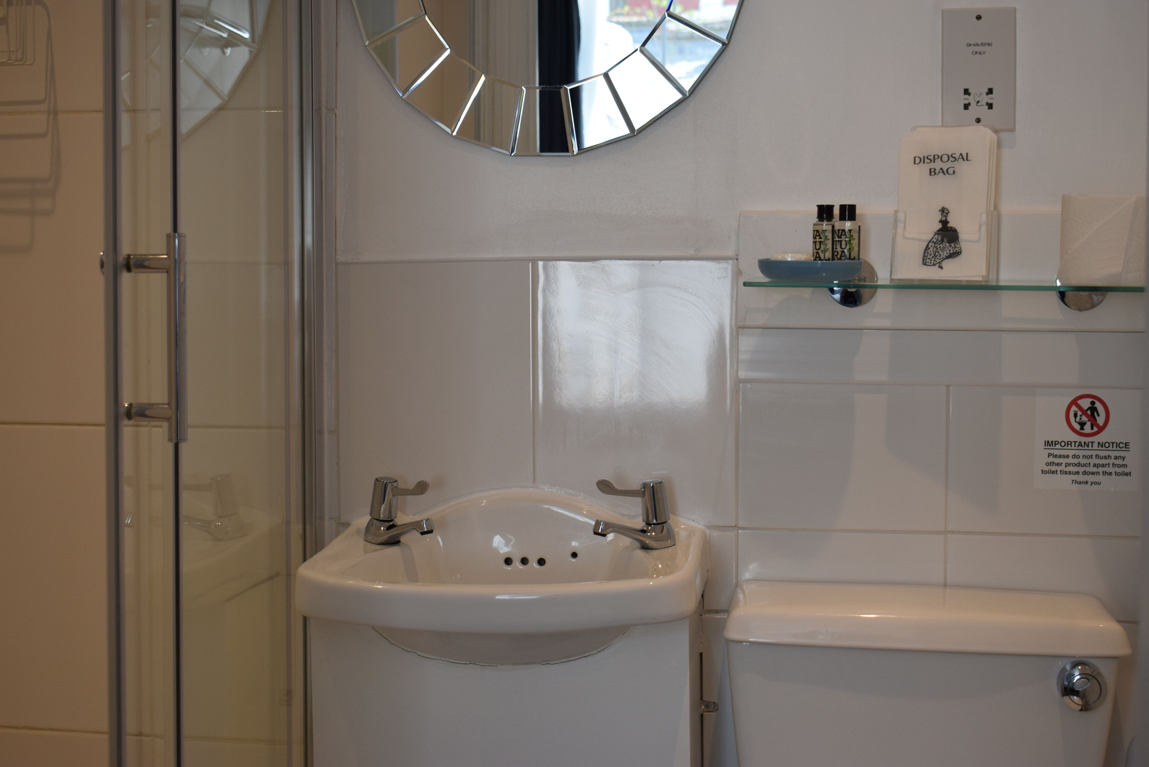 Un baño con lavamanos blanco, cabina de ducha con paneles de vidrio y una bolsa de residuos en un estante.