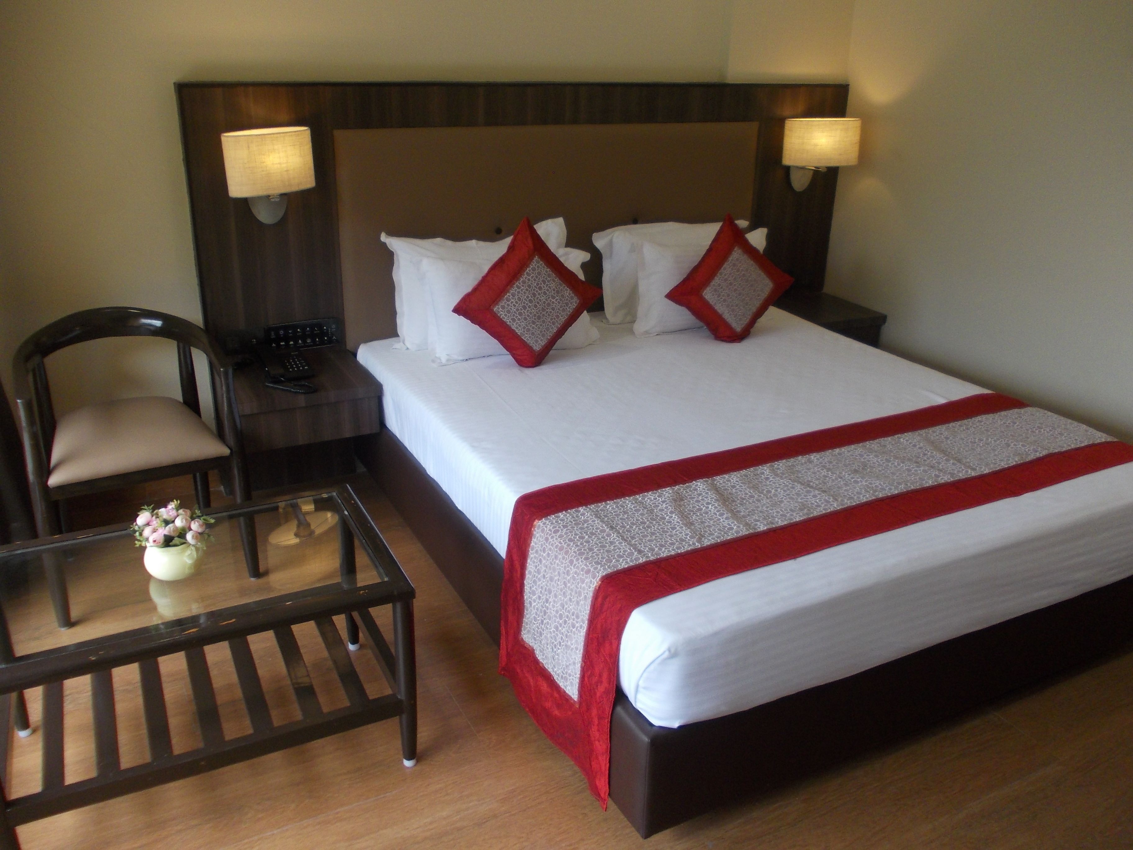 A hotel room with a bed, a chair, a nightstand, a lamp, and a small table with a vase.