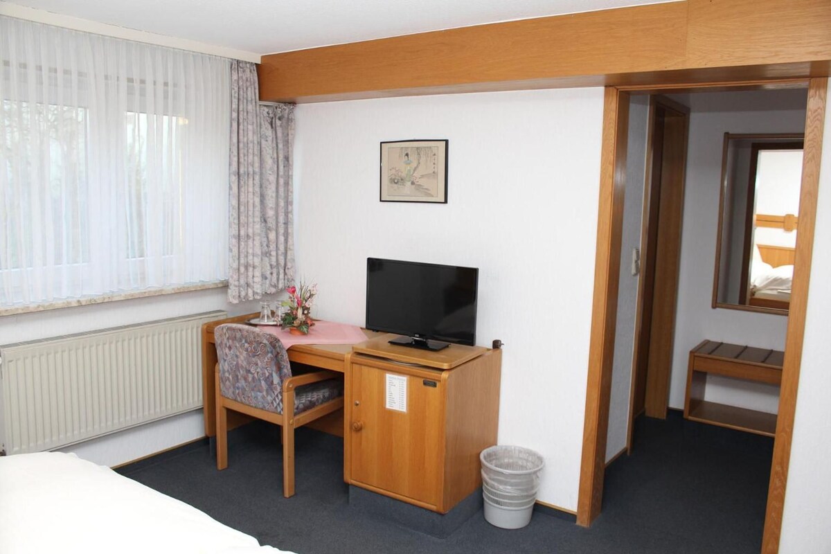 Une chambre d’hôtel avec un lit, un bureau avec un ordinateur, une chaise, un radiateur, une fenêtre avec des rideaux, un tableau au mur et une porte donnant sur une autre pièce.