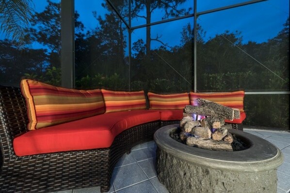 A wicker sofa with red cushions and striped pillows, a stone fire pit, and a view of trees through a glass wall.