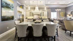 Un área de comedor moderna con una gran mesa rectangular rodeada de sillas estampadas, una cocina al fondo y un cuadro abstracto de gran tamaño en la pared.