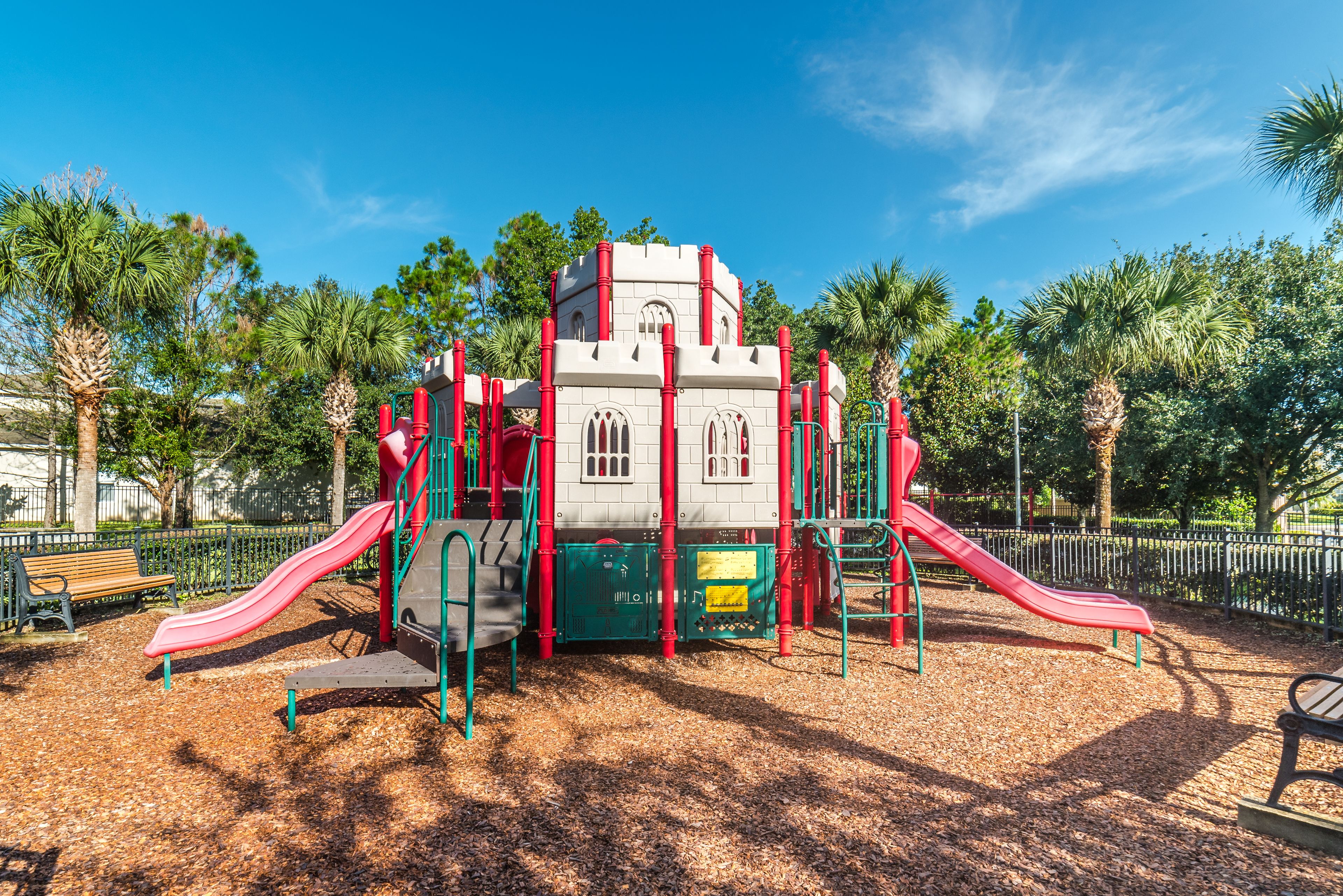 A playground with slides and climbing structures, surrounded by trees and a clear sky.