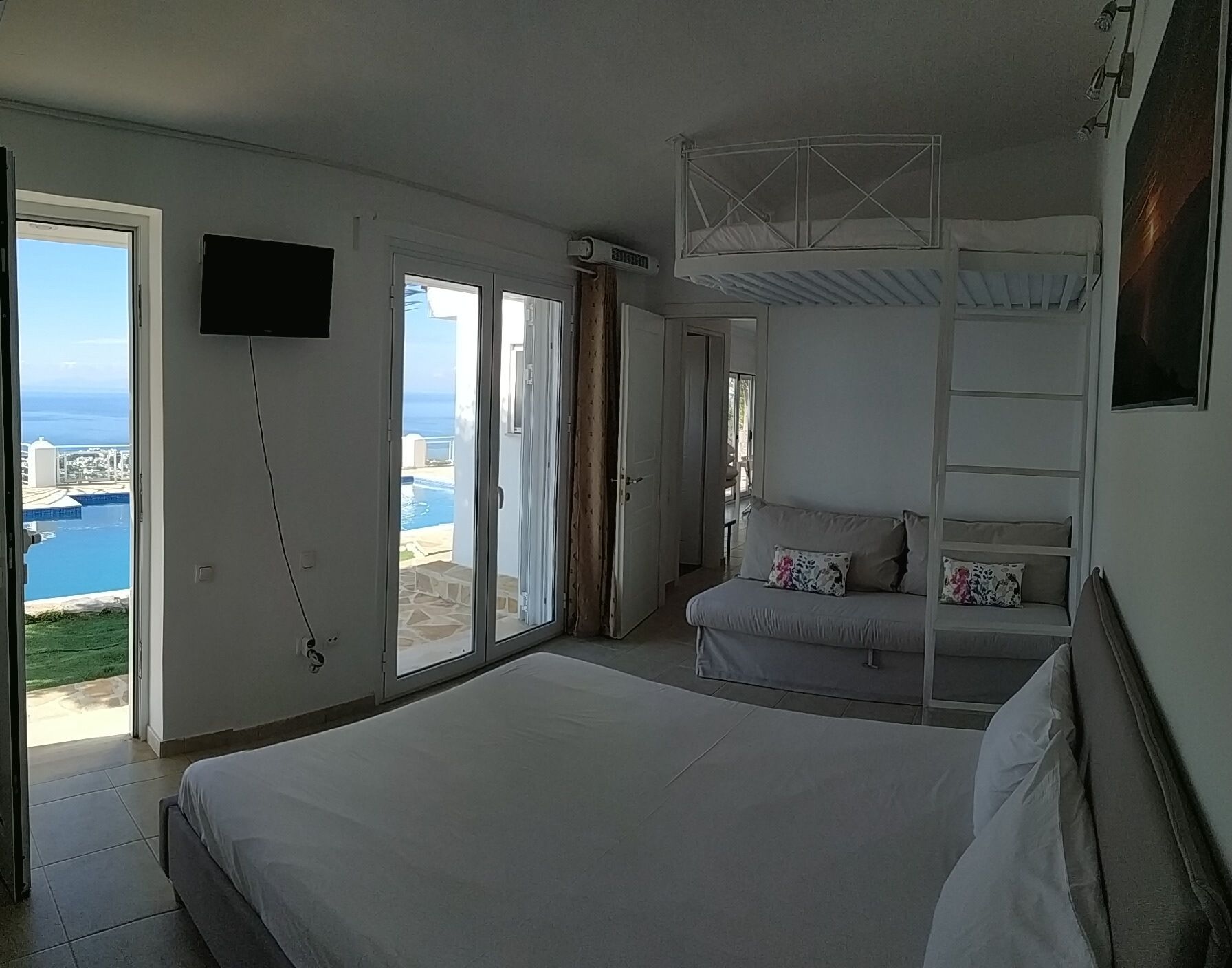 A bedroom with a large bed, a TV mounted on the wall, and a balcony with a pool view.