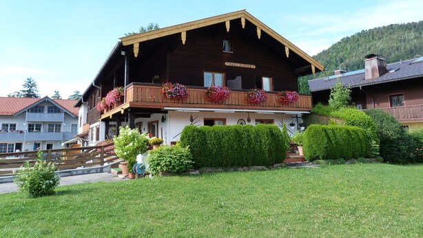 Un bâtiment traditionnel en bois, doté d’un balcon orné de fleurs, d’une enseigne indiquant « HAUS BERGER » et d’une pelouse bien entretenue à l’avant.