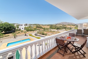 A balcony with a pool, dining table, and chairs, overlooking a landscape with buildings and hills.