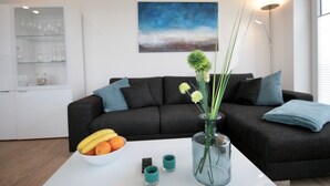 Un salon moderne avec un canapé gris foncé, une table basse blanche sur laquelle se trouve un saladier rempli de fruits, un vase avec des fleurs et un tableau accroché au mur.