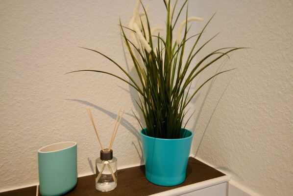A teal potted plant, a clear glass bottle with a brown cap, and a teal mug on a shelf against a white wall.