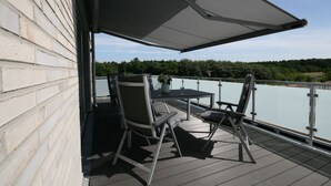 Une terrasse extérieure avec une table et des chaises, donnant sur une étendue d’eau et une zone verdoyante.