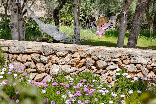 A hammock tied between trees, a stone wall, and a garden with flowers.