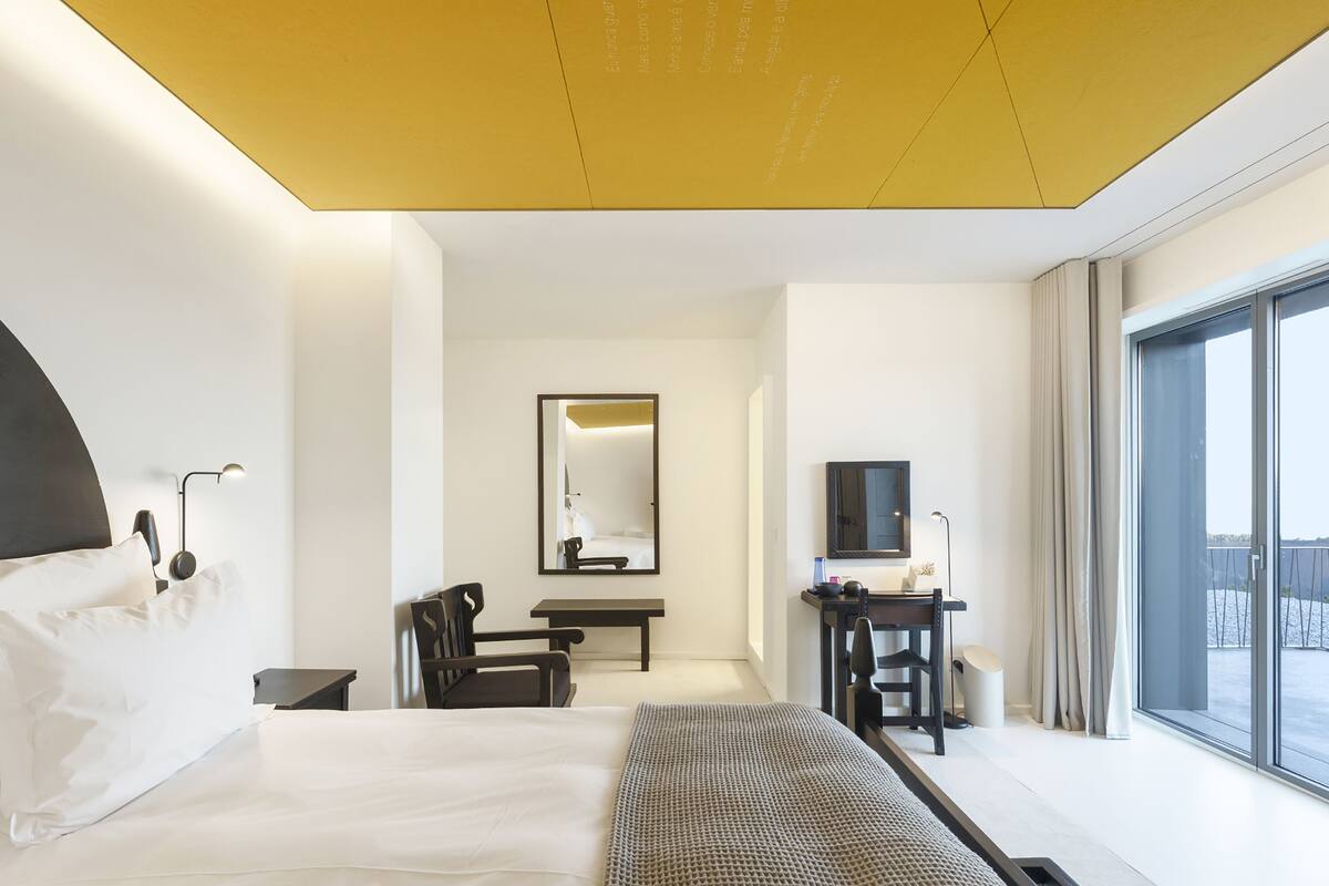 A modern hotel room with a large bed, a desk, a chair, a mirror, and a large window offering a view.