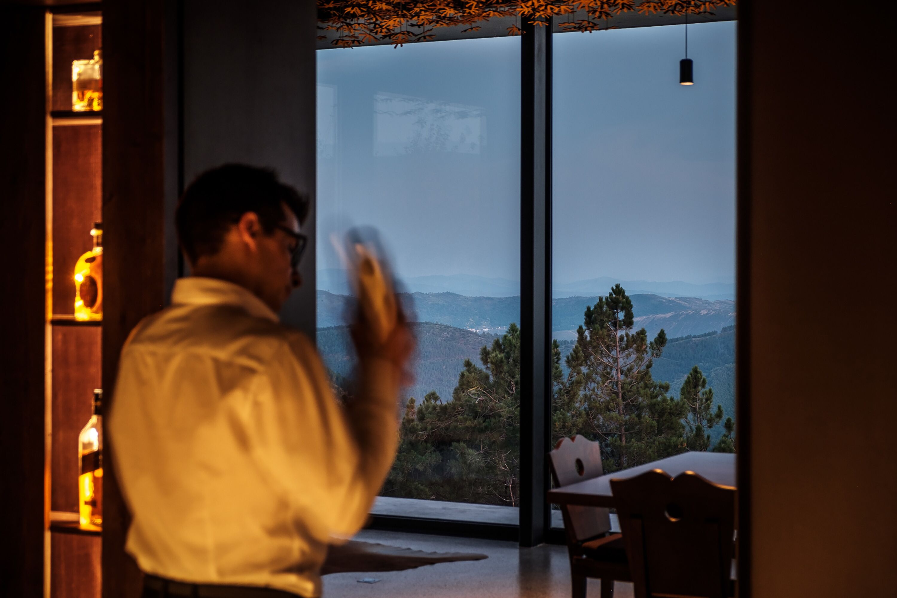 A man in a white shirt standing in a room with a large window overlooking a mountainous landscape.