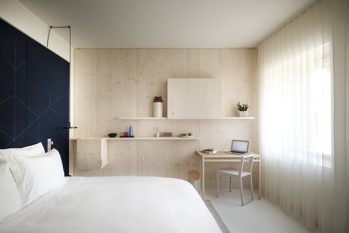 A modern bedroom with a bed, a desk, and a chair. There is a shelf above the desk with various items.