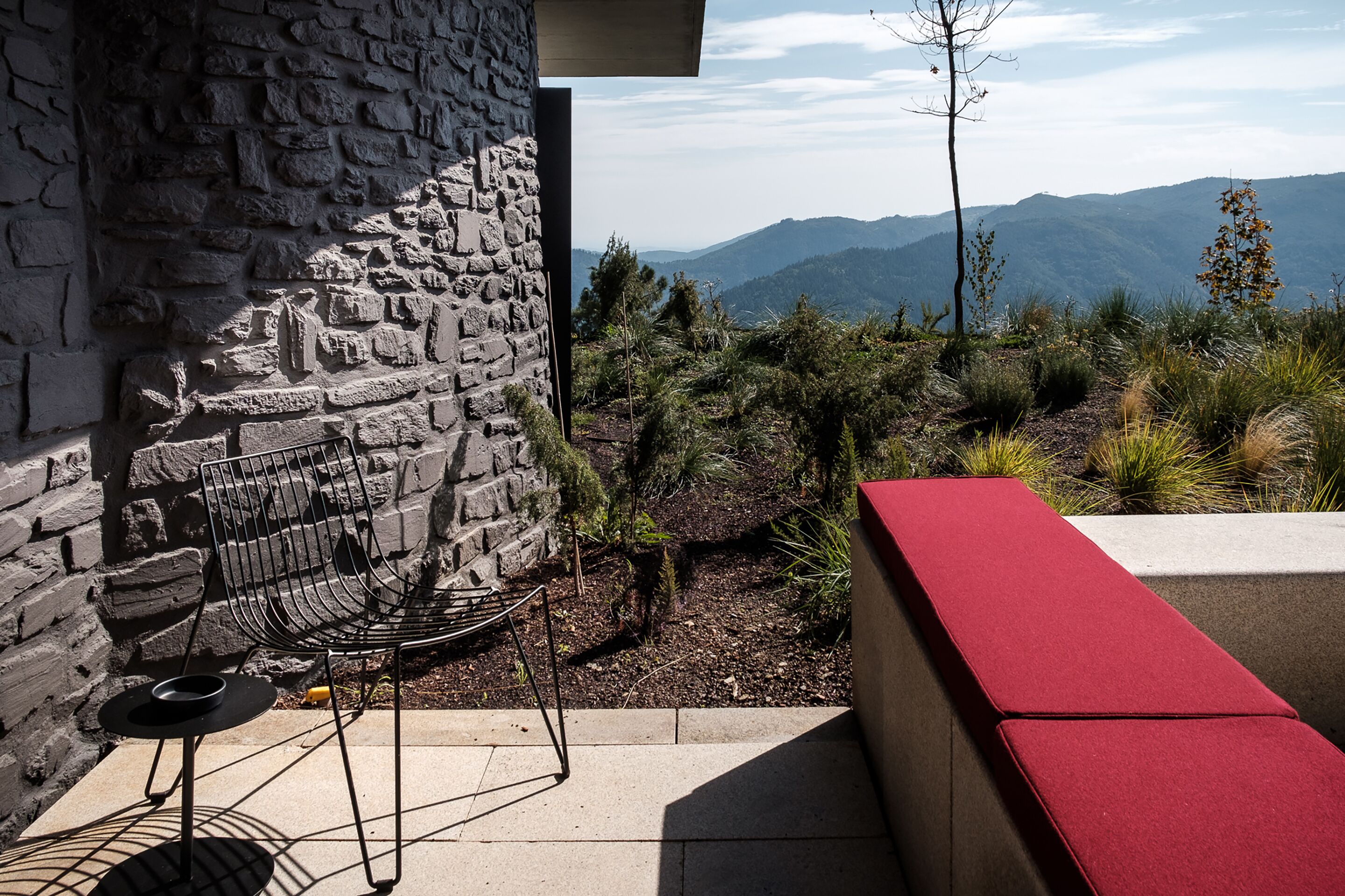 Un muro de piedra, una silla de metal y un banco rojo en un entorno exterior con vistas a la montaña.