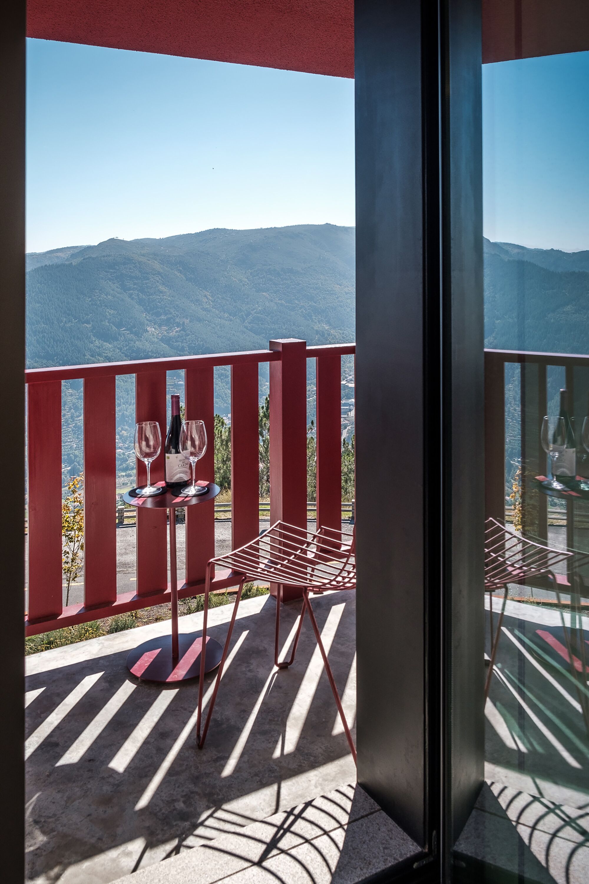 A balcony with a table set for two, a bottle of wine, and a glass. The balcony overlooks a mountainous landscape.