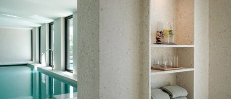Una piscina interior moderna con agua cristalina, una pared de concreto con repisas que sostienen toallas y vasos, y un ventanal grande que proporciona luz natural.