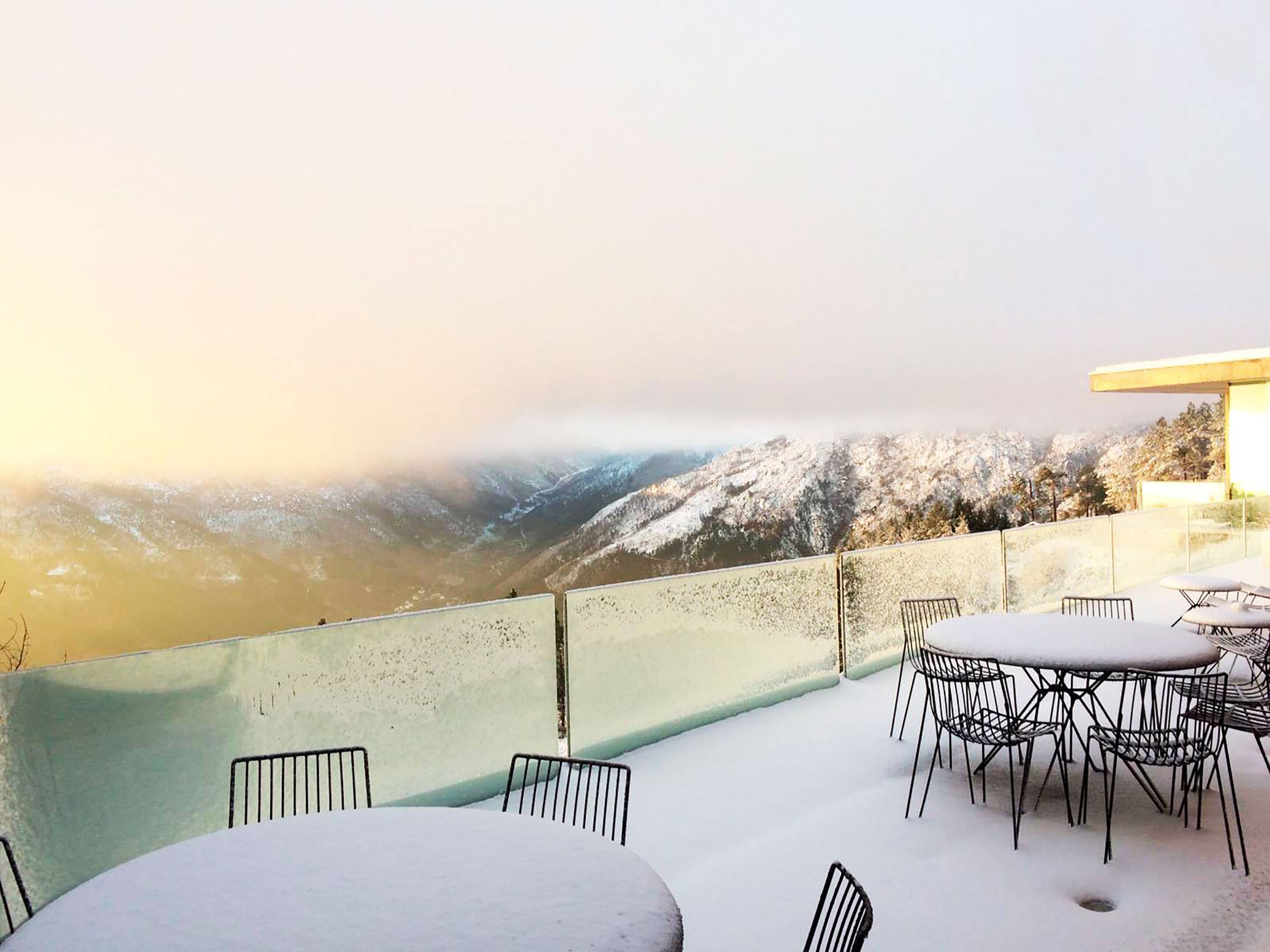 A snow-covered outdoor terrace with tables and chairs, overlooking a mountainous landscape.