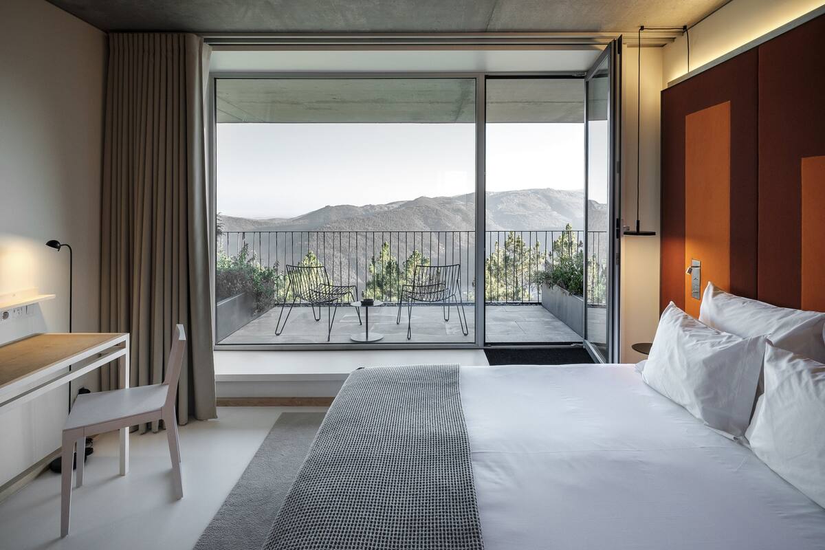 A modern hotel room with a large bed, a desk, and a balcony with a view of mountains.