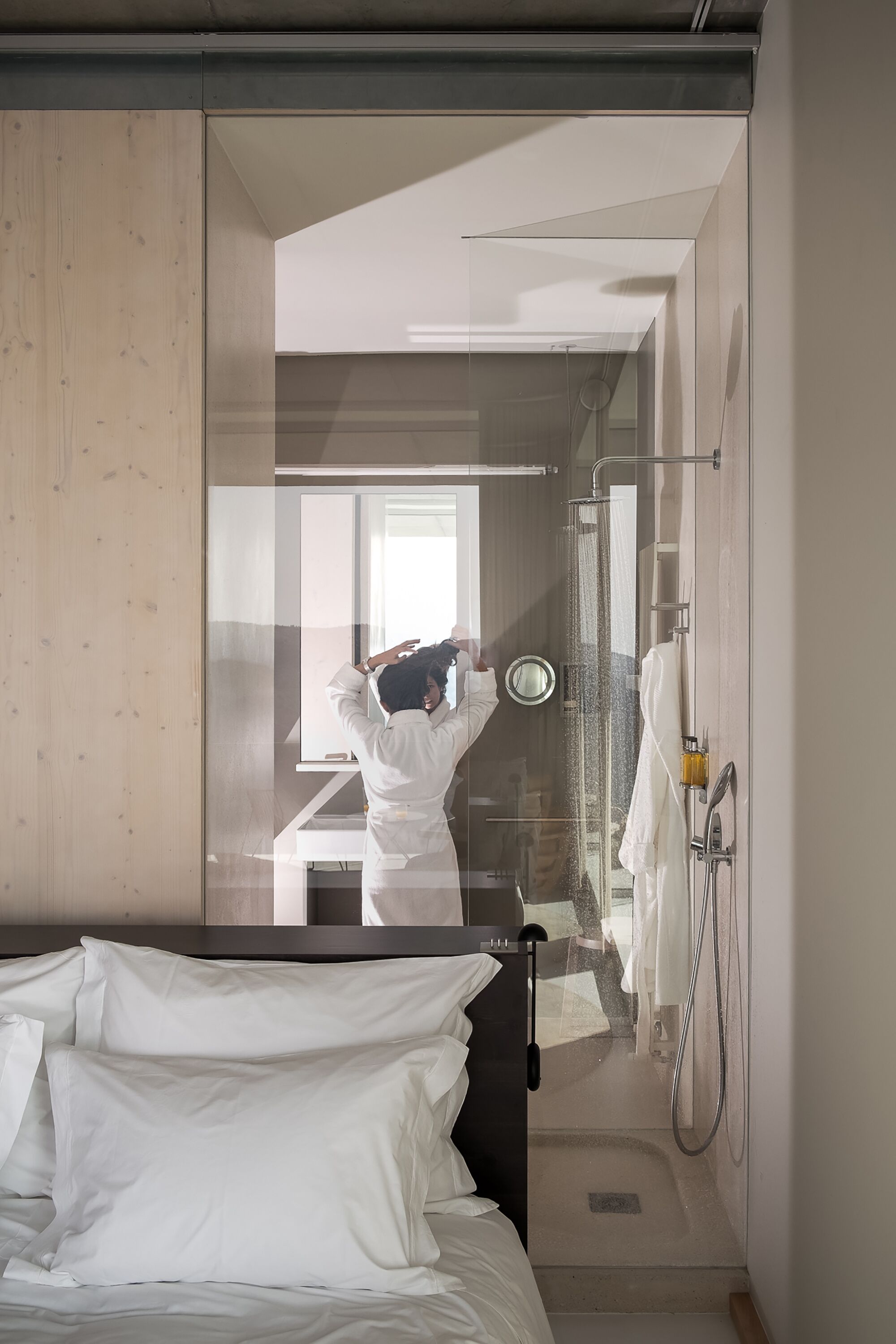 A modern hotel room with a glass partition, a bed with white linens, a shower area, and a visible bathroom.