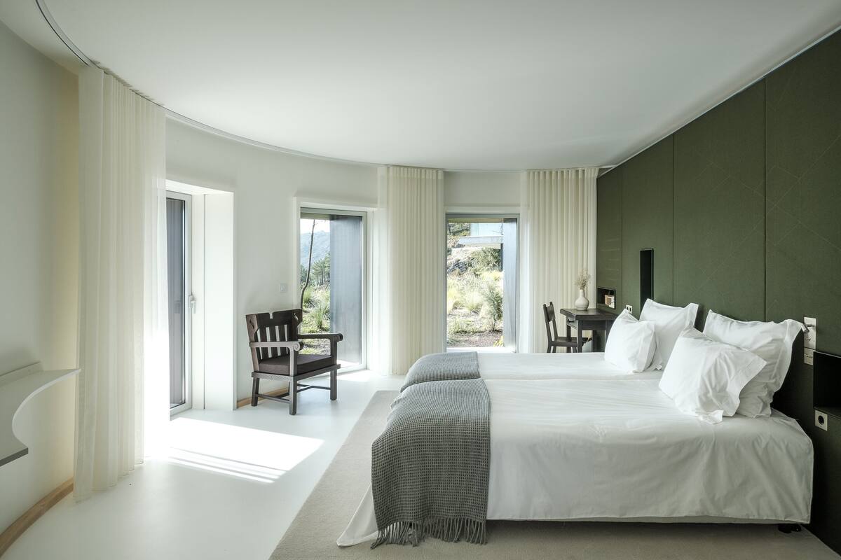 A modern bedroom with a large bed, a desk, and a view of the outdoors.
