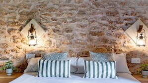 Un dormitorio con pared de piedra, dos mesitas de noche con lámparas, una cama con almohadas a rayas y una planta en maceta a cada lado.