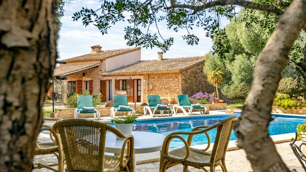 Zona junto a la piscina con sillas de mimbre y un edificio de piedra al fondo.