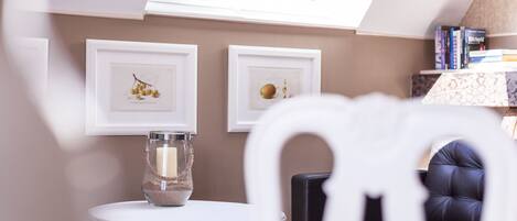 A cozy room with a skylight, framed botanical prints on the wall, a glass candle holder on a white table, and a dark leather chair.