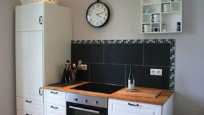 A kitchen with a black tiled backsplash, a wooden countertop, a white cabinet, a clock, and a shelf with cups.