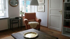 A room with a wooden floor, a round table with a pie on it, a chair with a cushion, a lamp, and a door with a shelf above it.
