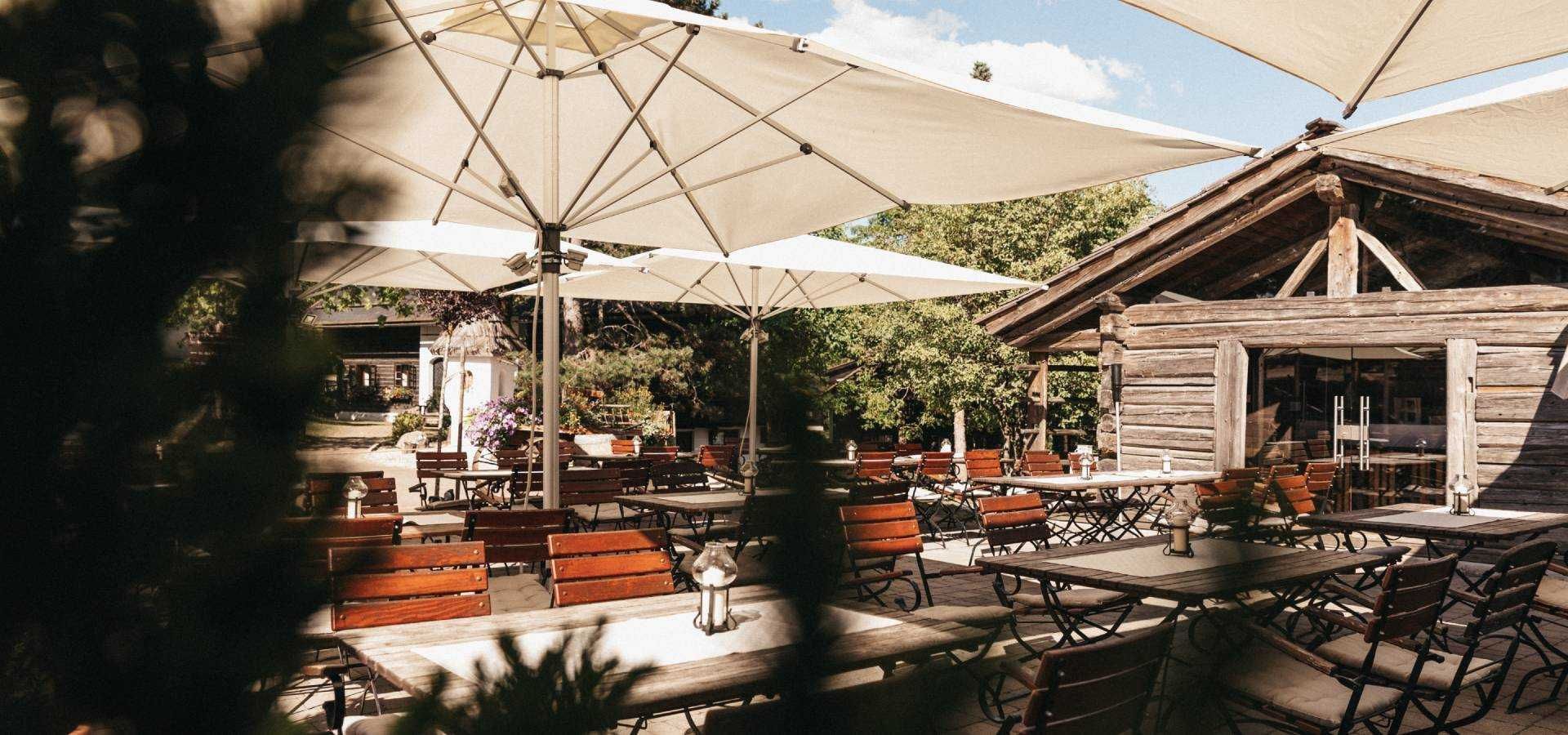 An outdoor patio with wooden tables and benches, shaded by large umbrellas, and a rustic wooden building in the background.