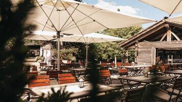 An outdoor patio with wooden tables and benches, shaded by large umbrellas, and a rustic wooden building in the background.