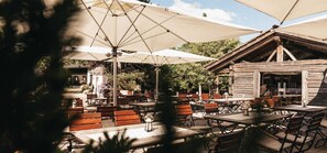 Une terrasse extérieure avec des tables et des bancs en bois, ombragée par de grands parasols, et un bâtiment rustique en bois en arrière-plan.