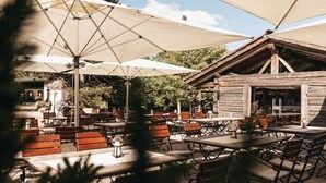 Un patio al aire libre con mesas y bancos de madera, cubierto por grandes sombrillas, y un rústico edificio de madera al fondo.