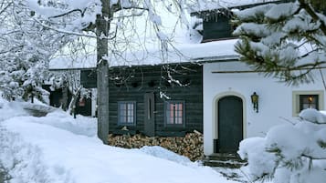 A snow-covered wooden house with a dark door and windows, a small porch, and a visible chimney.