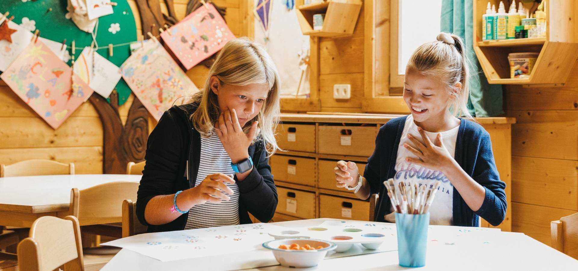 Deux personnes sont assises à une table, entourées de matériel de dessin, de meubles en bois et, au fond, d’un tableau d ’affichage.