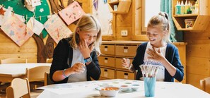 Two people at a table with art supplies, wooden cabinets, and a bulletin board in the background. - Apartment Schwindelfrei - Naturel Hoteldorf SCHÖNLEITN (Latschach am Faaker See)