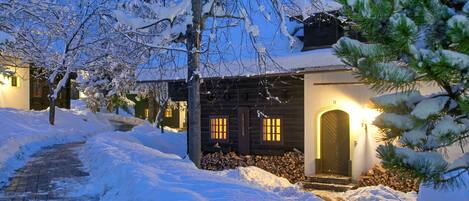 Ein verschneiter Pfad führt zu einer Holzhütte mit beleuchtetem Eingang und Fenstern.