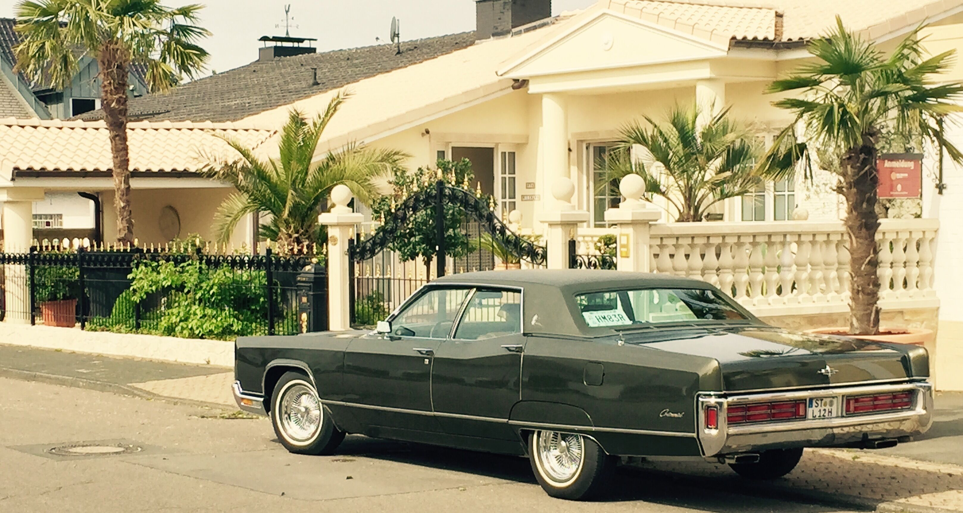 Un coche negro clásico estacionado frente a una casa de fachada blanca, una cerca negra y palmeras.