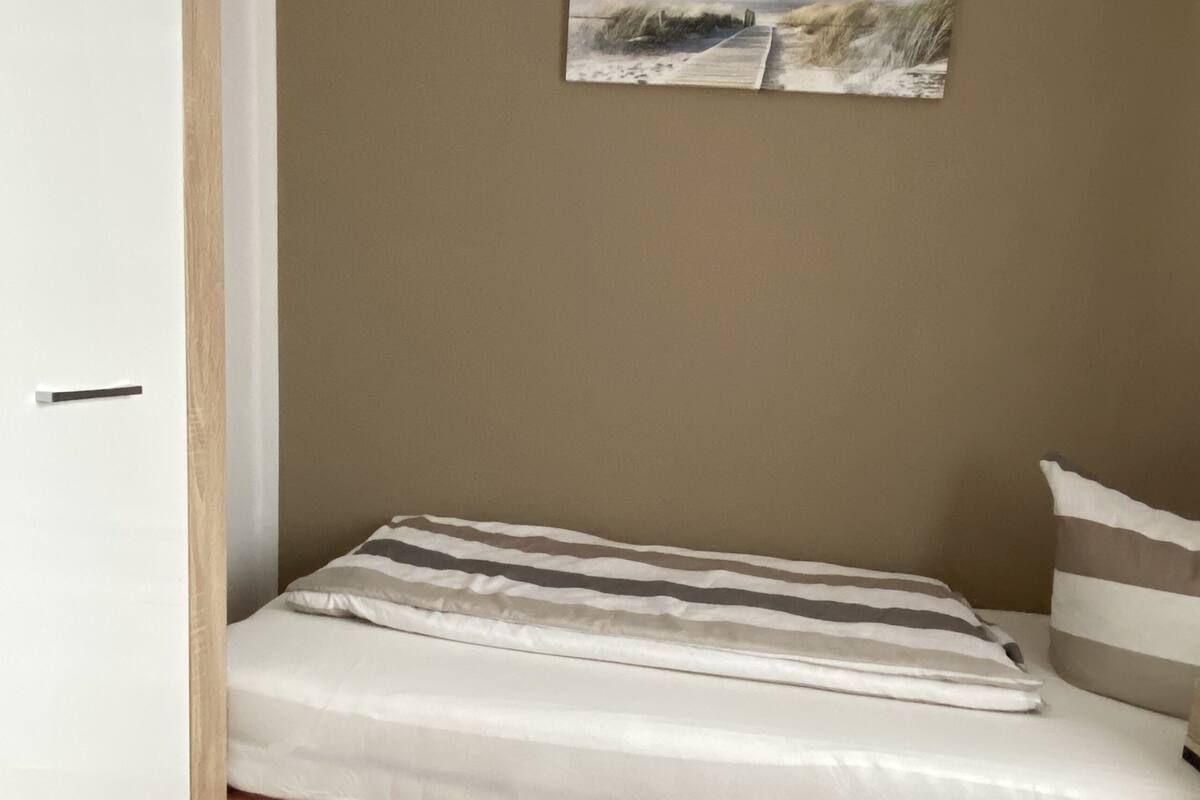 Quarto pequeno com uma cama de solteiro, piso de madeira, parede bege e um quadro pendurado na parede.