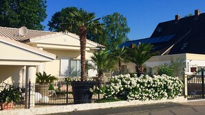A residential building with a sloped roof, a gated entrance, and a landscaped garden with white flowers and palm trees.