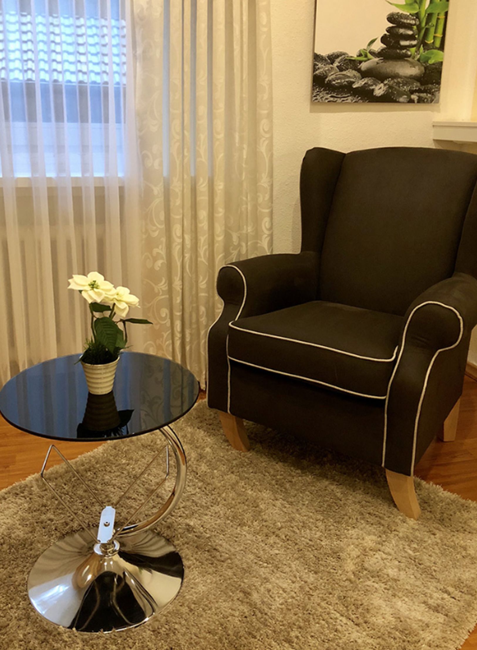 Un salon avec un fauteuil de couleur marron foncé, une petite table ronde ornée d’un vase de fleurs blanches et un tableau représentant des rochers accroché au mur.