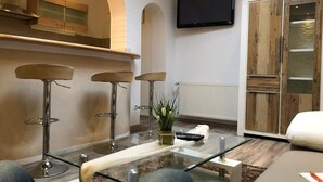 A modern living room with a glass coffee table, three bar stools, a television mounted on the wall, and a sliding wooden door.