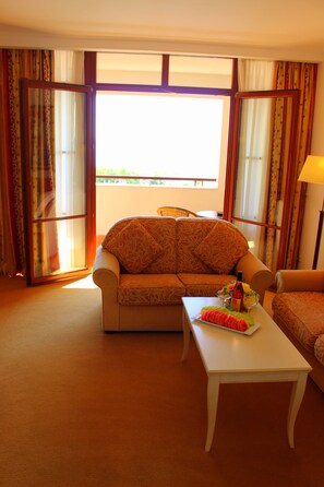A hotel room with a sofa, a small table with a fruit bowl, and a view through a large window. - Helena Sands (Nessebar)