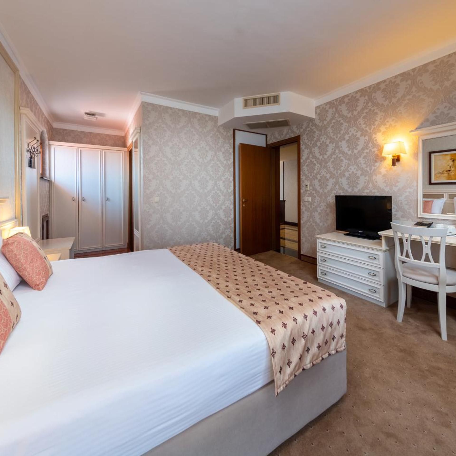 A hotel room with a bed, a desk with a chair, a television, a wardrobe, and a mirror.