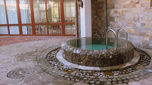 A stone hot tub with metal ladders, surrounded by a stone mosaic floor, near a glass door with a wooden frame. - Helena Sands (Nessebar)