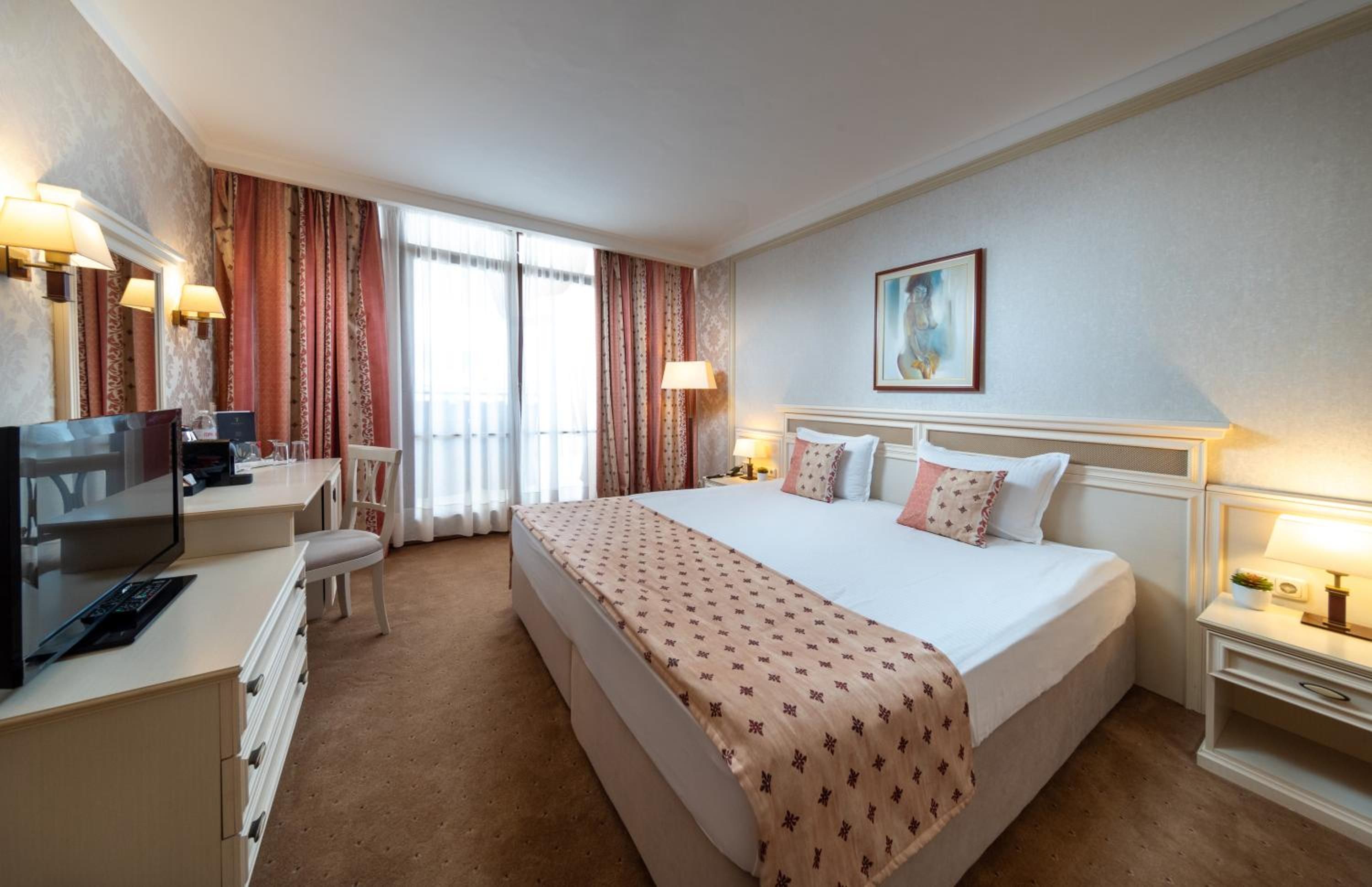 A hotel room with a bed, desk, chair, TV, and a painting on the wall.